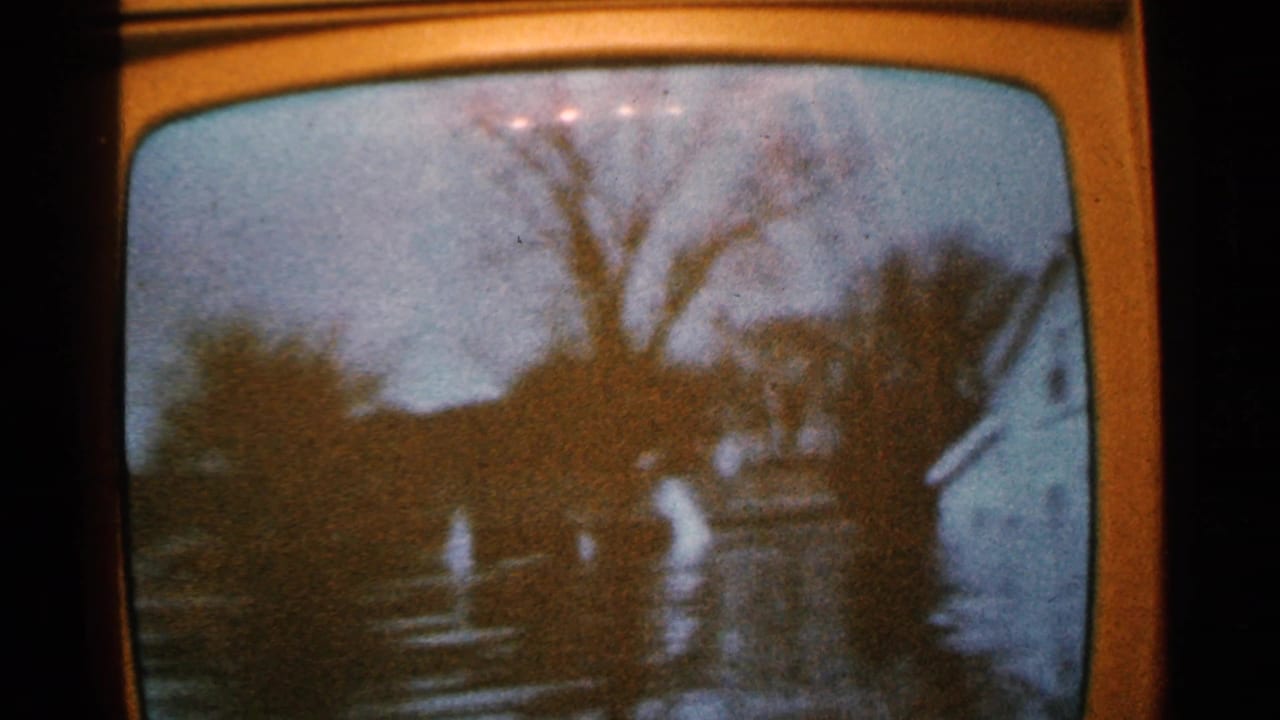 A person in light clothing stands or walks in floodwater near houses and bare trees.