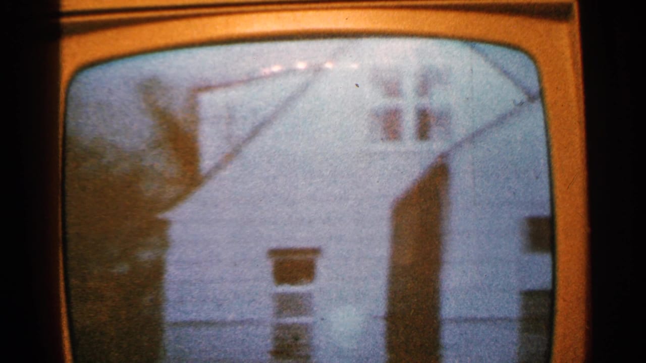 1965 Minnesota 8mm frame of a rounded television screen showing a soft, grainy view of a white house exterior with dark rooflines and bare trees.