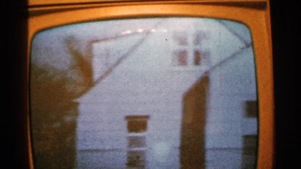 1965 Minnesota 8mm frame of a rounded television screen showing a soft, grainy view of a white house exterior with dark rooflines and bare trees.