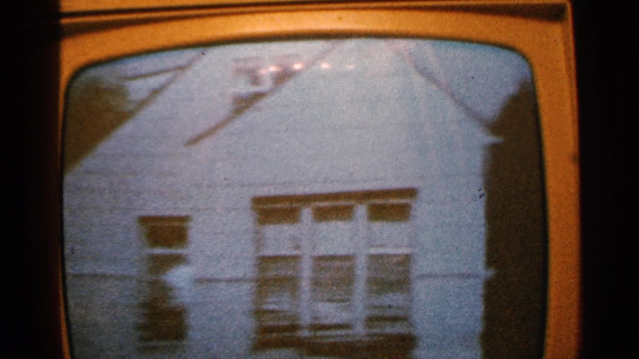 1965 Minnesota home-movie frame showing a white house facade on an old television screen, with visible roofline, windows, and screen curvature.