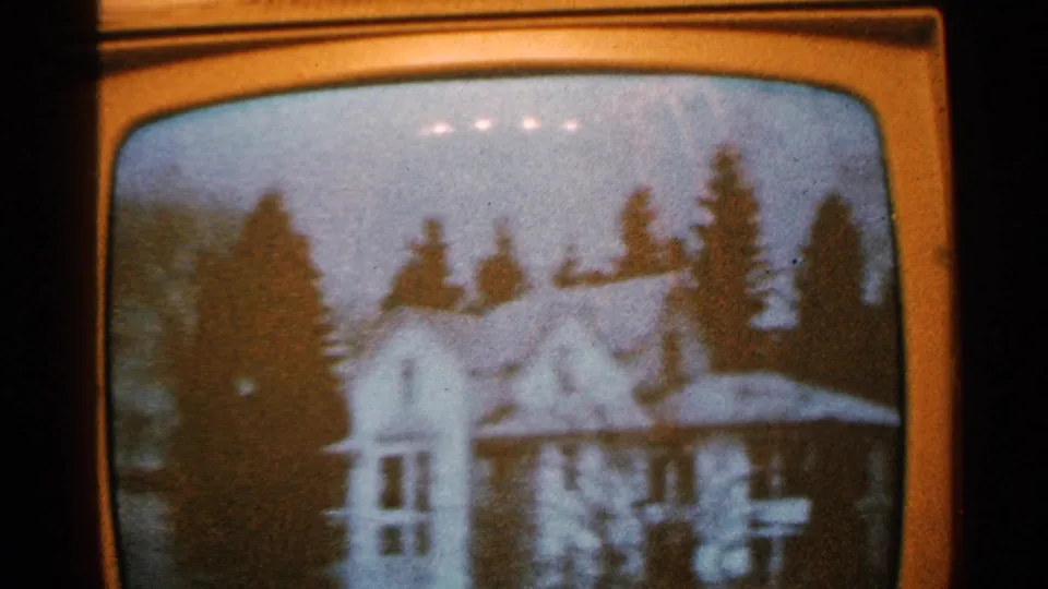 1965 Minnesota frame showing a wider television-screen view of a white residential house surrounded by tall trees and pale winterlike ground or roof surfaces.