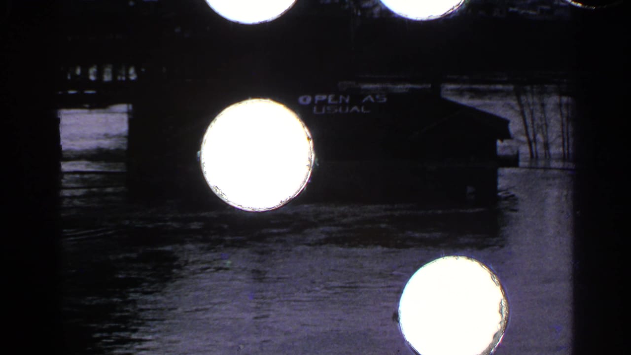 Floodwater surrounds a dark riverside building with bright film flares over the frame.