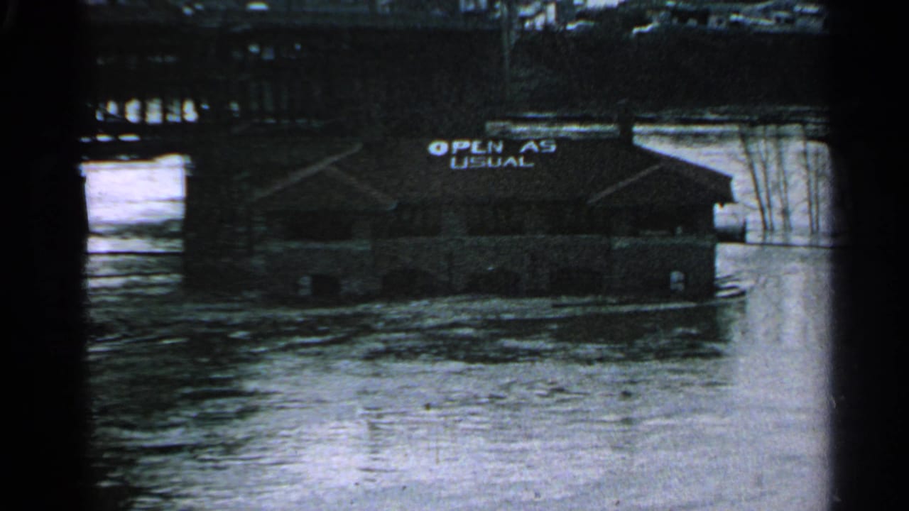 Minnesota 1965 flood footage shows a low riverside building with an 'OPEN AS USUAL' sign above moving water.