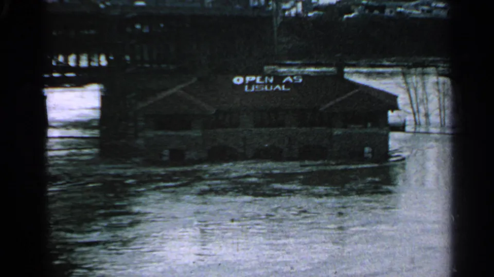 Minnesota 1965 flood footage shows a low riverside building with an 'OPEN AS USUAL' sign above moving water.