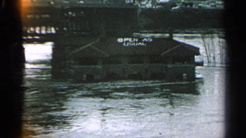 Minnesota 1965 8mm frame of a partially surrounded riverside building, readable 'OPEN AS USUAL' sign, and churning floodwater.