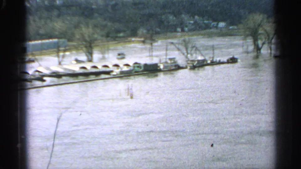 Minnesota 1965 flood scene showing a long dock or barge line on high water with bare trees and a pale flooded bank.