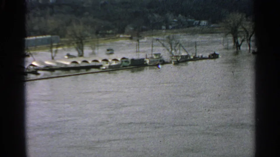 1965 Minnesota 8mm frame showing high floodwater, bare trees, and a line of boats or barges near the far bank.