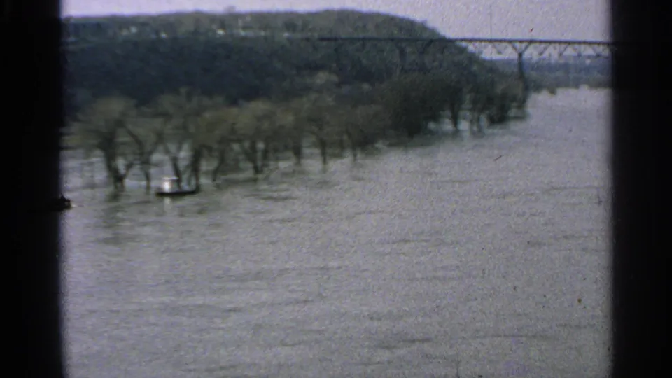 1965 Minnesota flood scene showing submerged riverside trees, broad water, a distant bridge, and a dark wooded ridge.