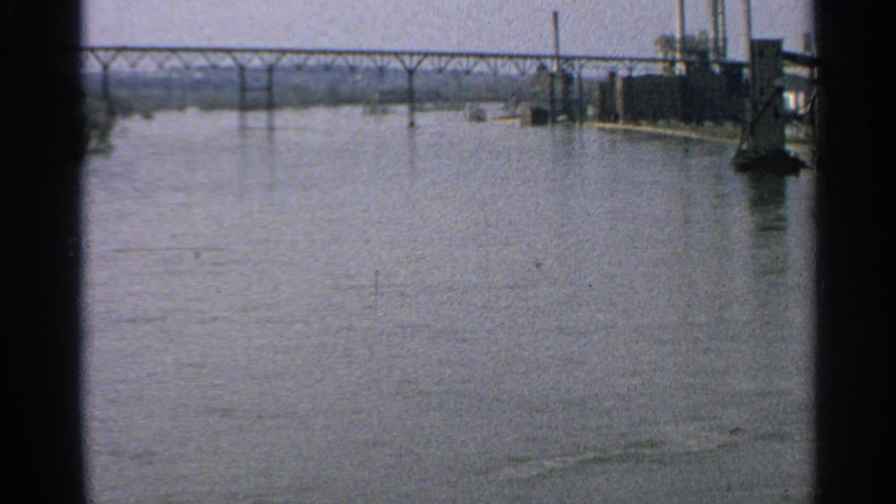 1965 Minnesota river scene with a distant steel bridge and industrial waterfront structures visible at the right edge.