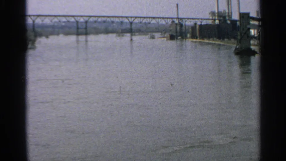 1965 Minnesota river scene with a distant steel bridge and industrial waterfront structures visible at the right edge.
