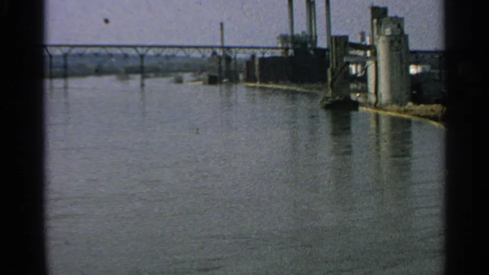 1965 Minnesota 8mm frame showing high river water beside an industrial waterfront with towers and a bridge beyond.