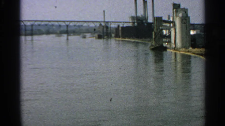 1965 Minnesota riverfront view with industrial structures, a distant bridge, and high water filling the foreground.