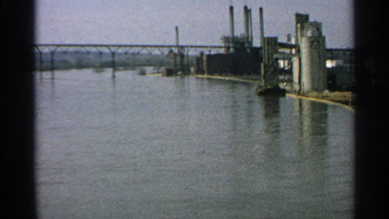 Wide river view with a bridge, industrial buildings, silos, and smokestacks along the shoreline.
