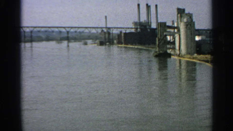 1965 Minnesota 8mm view of a flooded riverfront with a bridge, factory structures, vertical silos, and smokestacks near the water.