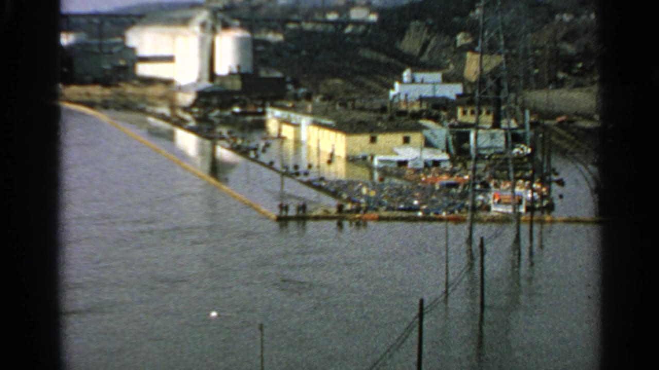 Flooded waterfront with industrial buildings, parked cars, utility poles, and a yellow floating barrier.