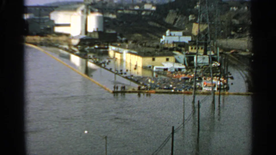 1965 Minnesota home-movie frame showing floodwater beside a low industrial waterfront, with parked cars and utility poles near the river edge.