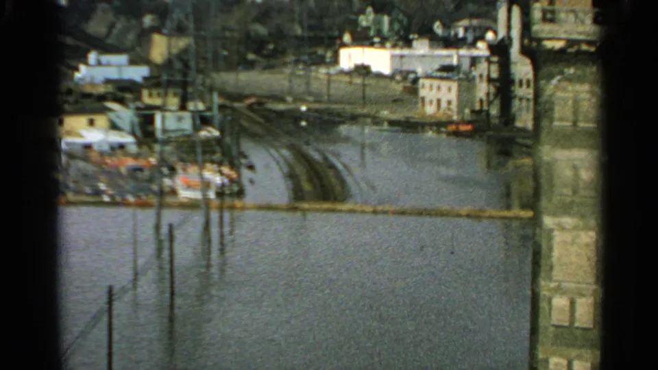 1965 Minnesota 8mm frame of an industrial riverfront where high water reaches tracks or roads near buildings, poles, and a smokestack.