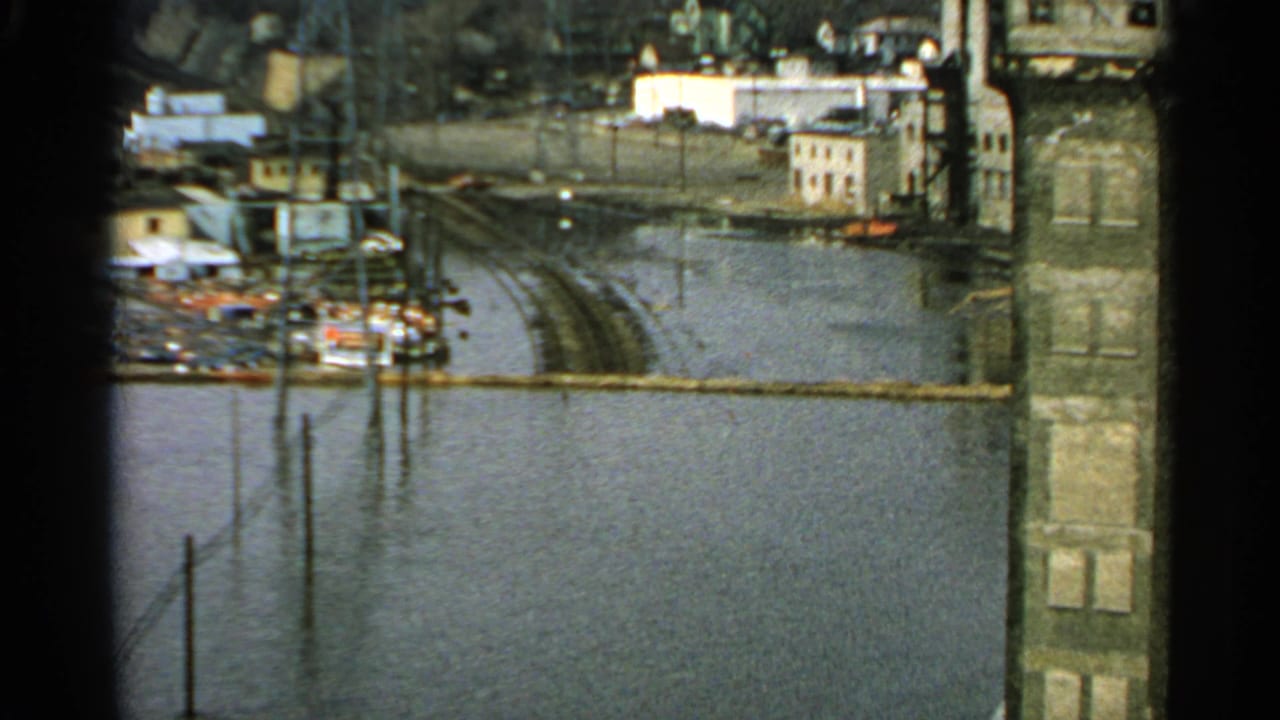 1965 Minnesota 8mm frame showing floodwater around buildings, poles, and a submerged roadway or rail line near industrial structures.