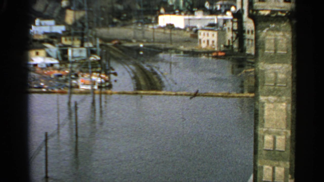 1965 Minnesota flood scene with water covering low ground, buildings at the edge, utility poles, and industrial structures in the distance.