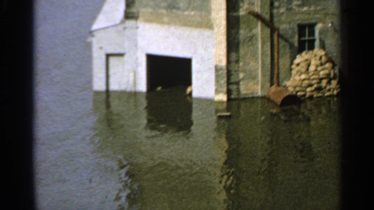 Minnesota industrial flood scene from 1965 showing water at factory doorways, sandbags against a wall, and a barrel near the flooded entrance.