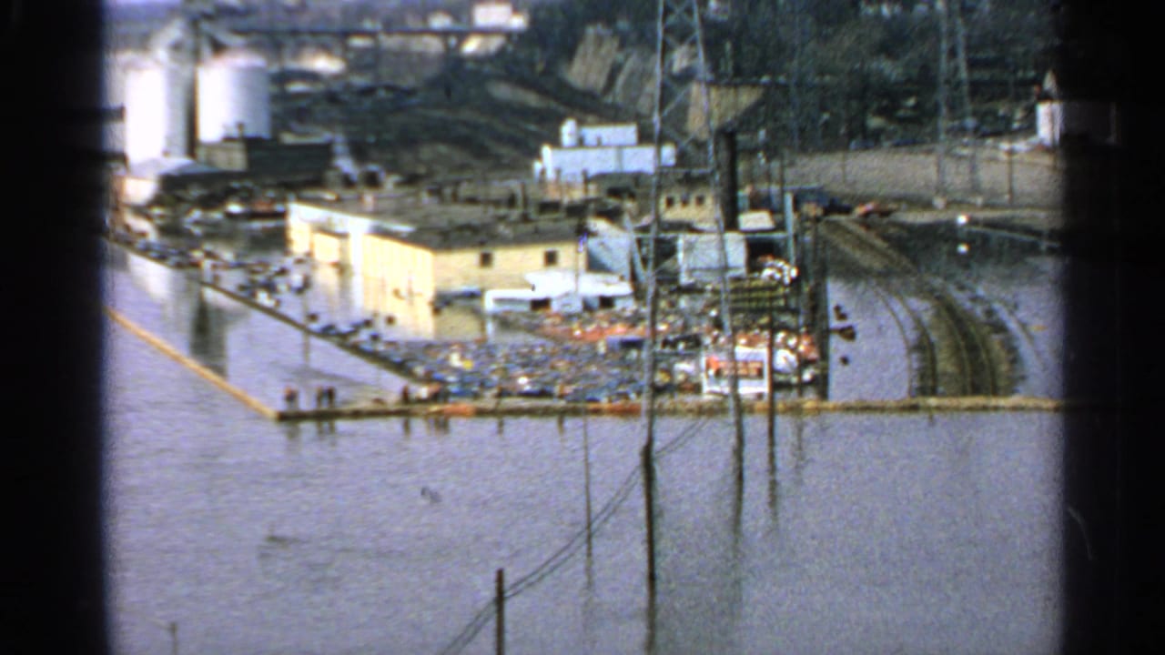 Wide flooded industrial yard with buildings, rail lines, utility poles, tanks, and debris.