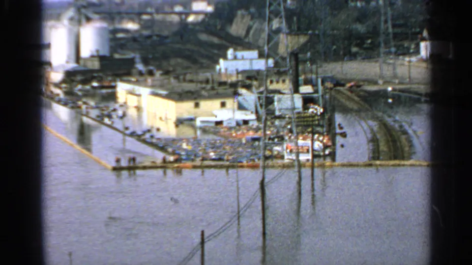 Wide 1965 Minnesota view of an industrial complex surrounded by floodwater, with submerged rail lines, poles, tanks, and scattered materials.