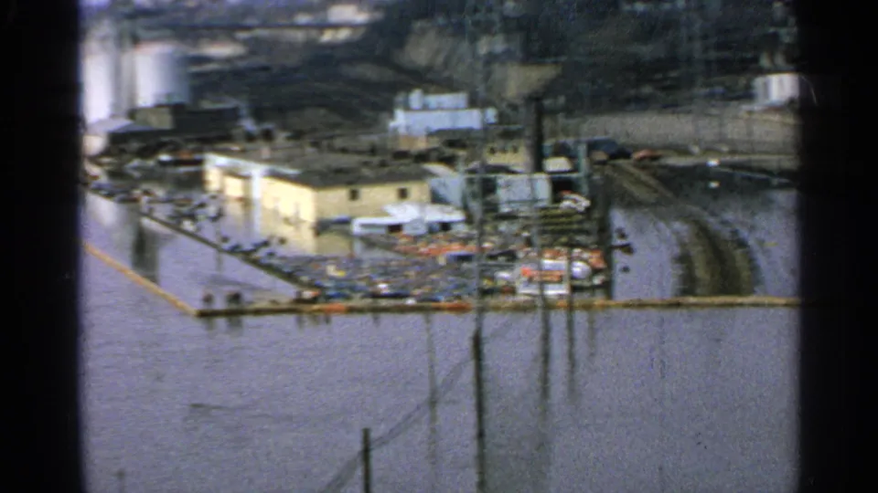 Elevated 1965 Minnesota shot of a flooded industrial complex, showing water around rail tracks, utility poles, buildings, and yard debris.