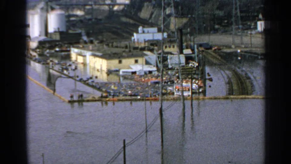1965 Minnesota flood view of factory grounds with rail lines, poles, tanks, and low industrial buildings surrounded by murky water.