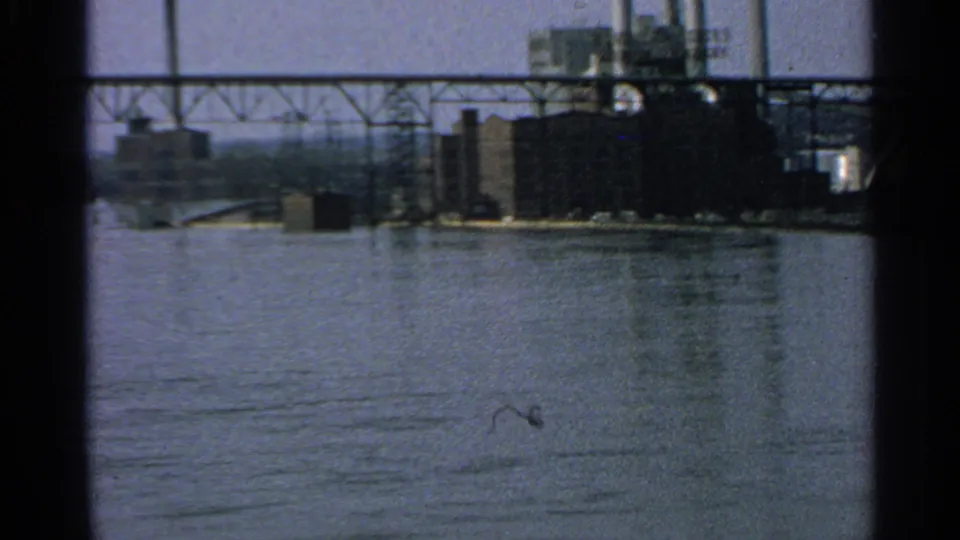 1965 Minnesota 8mm frame showing industrial buildings, smokestacks, and a bridge viewed across a river.