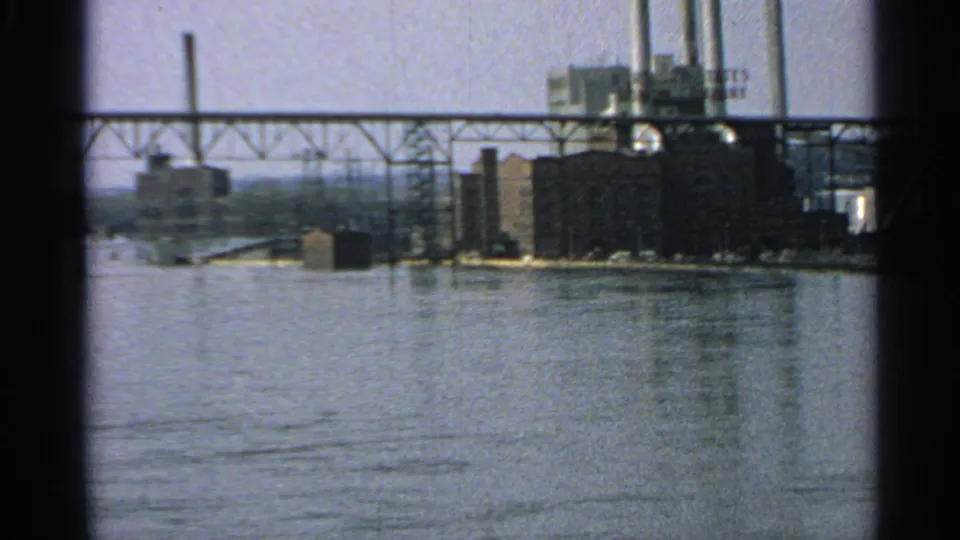 Faded 8mm view of a Minnesota industrial shoreline, showing factory structures, smokestacks, bridgework, and open water.