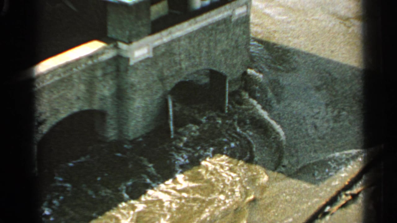 1965 Minnesota 8mm frame showing turbulent floodwater pushing through arched bridge openings from an elevated viewpoint.