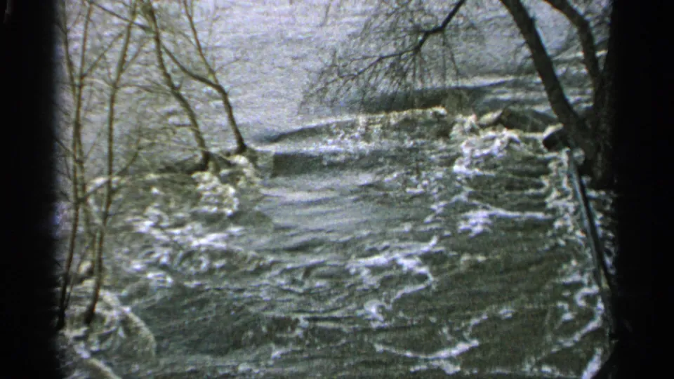 1965 Minnesota 8mm frame looking along a swollen river channel with bare branches, banks, and choppy floodwater.