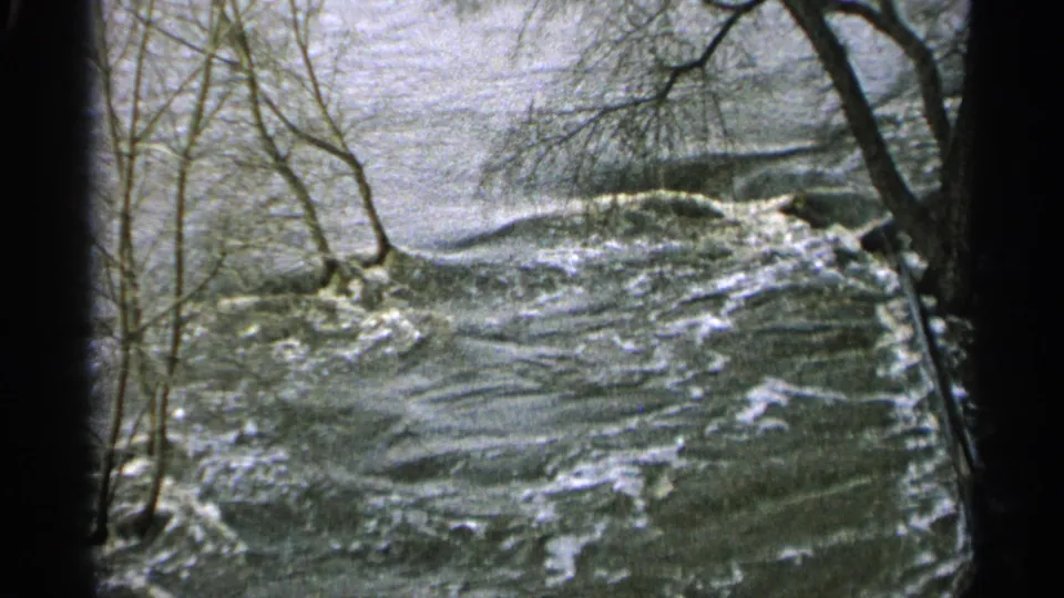 1965 Minnesota home-movie frame showing turbulent floodwater rushing past bare riverside trees.