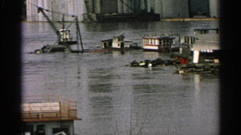 1965 Minnesota home-movie frame showing small boats, a crane, and waterfront buildings surrounded by floodwater.