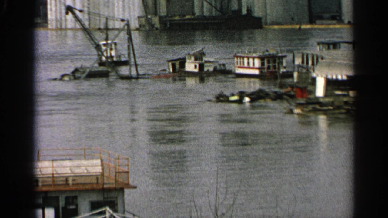 Flooded waterfront worksite with barges, a crane, debris, and industrial buildings across the water.
