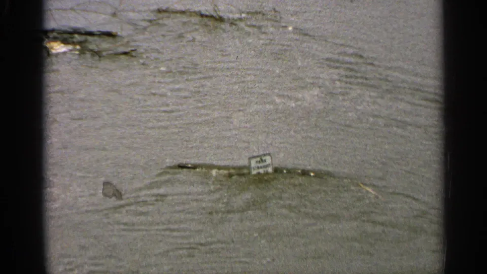Minnesota 1965 8mm frame focused on floodwater, ripples, floating debris, and a small sign partly surrounded by water.
