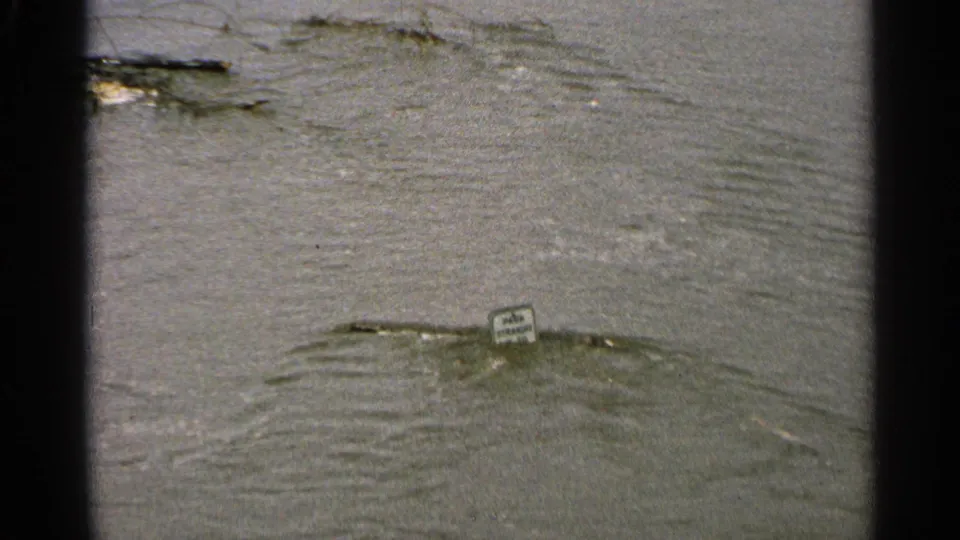 Minnesota 1965 8mm frame showing a small sign amid high water, with ripples and debris suggesting a flooded worksite edge.