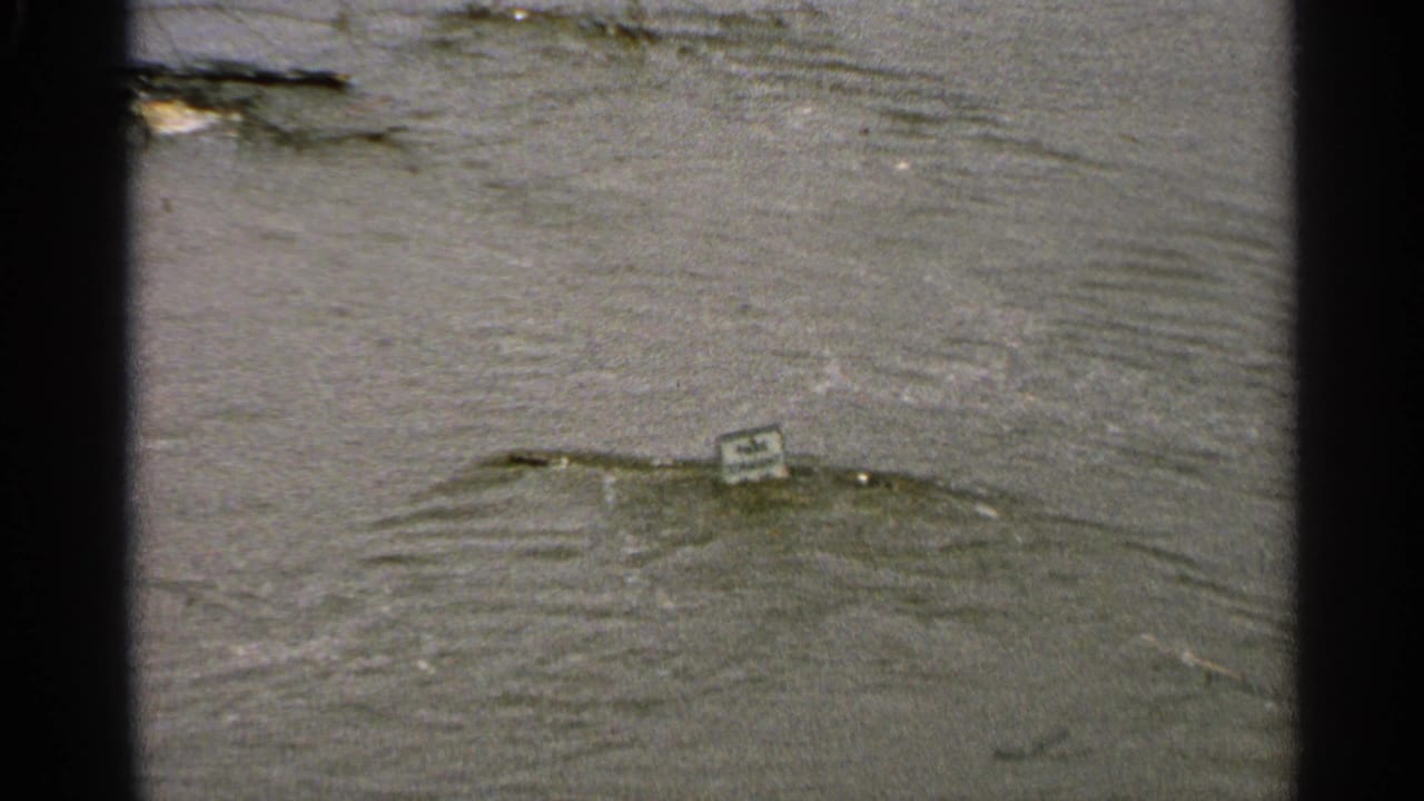 1965 Minnesota 8mm frame showing murky floodwater with ripples, debris, and a small sign partly submerged near the water surface.