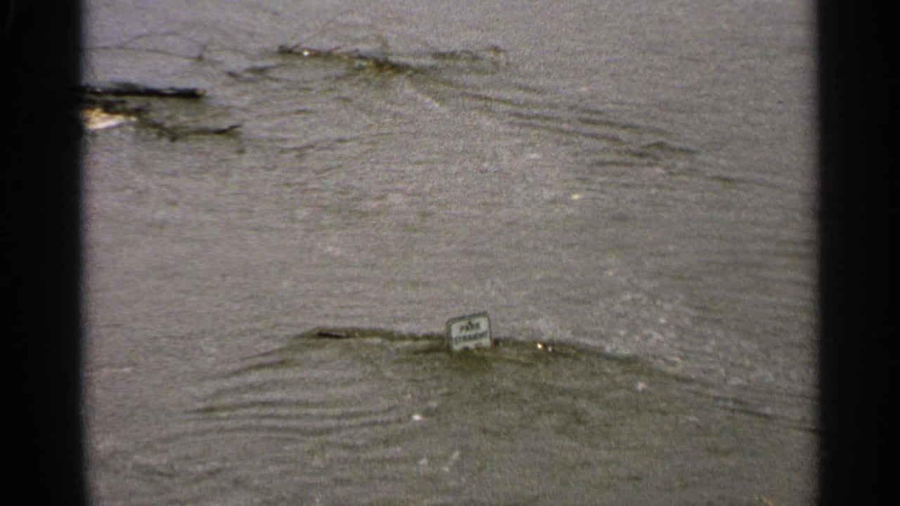 Floodwater surrounds a small sign with debris floating nearby.
