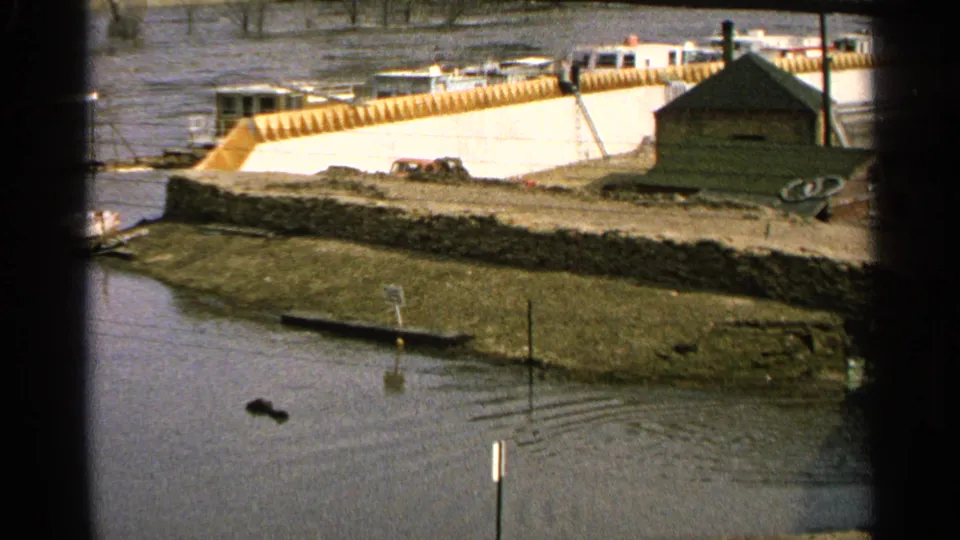 1965 Minnesota 8mm frame showing a wider flooded riverbank with a stone embankment, small shed, parked cars, industrial structures, and a submerged sign.