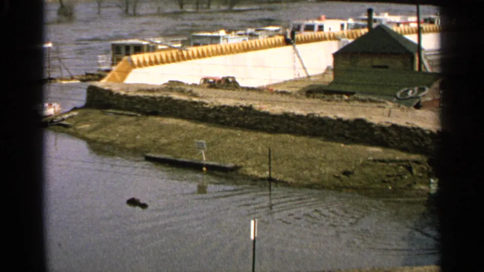1965 Minnesota 8mm frame showing floodwater along a river edge, a stone embankment, a small shed, parked cars, a submerged sign, and floating debris.
