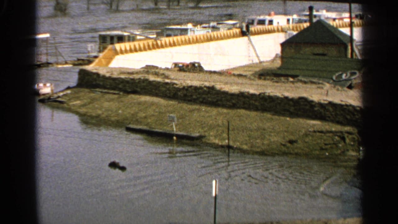 1965 Minnesota 8mm frame showing a flooded marina area with submerged dock edges, riverside structures, and boats seen through a dark foreground frame.