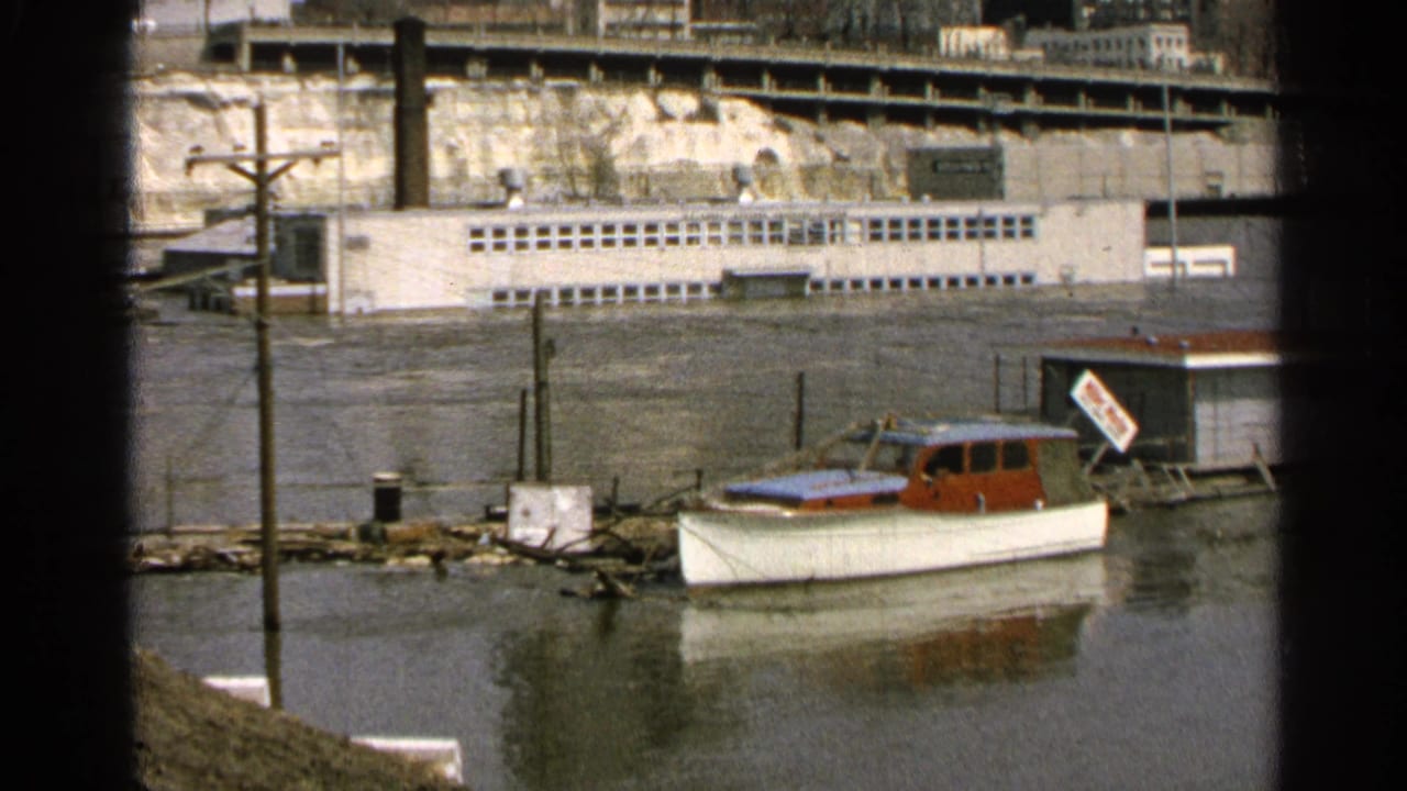 1965 Minnesota waterfront frame with a small white and red cabin boat moored beside partially submerged posts and a long building beyond the water.
