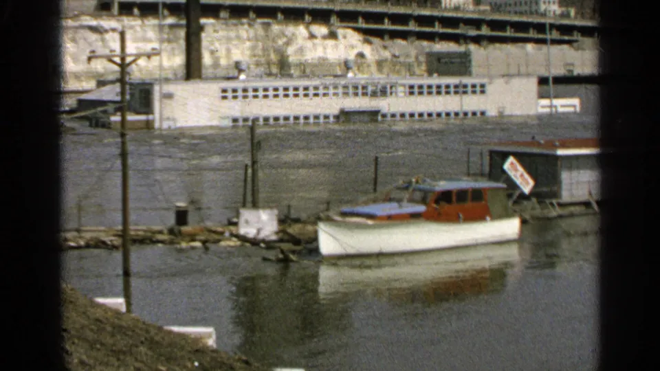 1965 Minnesota 8mm frame showing a flooded marina scene with a moored cabin boat, submerged shoreline, dock posts, and a long white building.