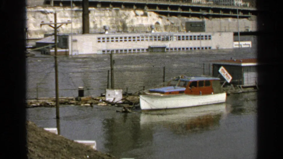 1965 Minnesota flooded marina frame with a small white and red boat near partially submerged posts, backed by a riverside building and retaining wall.