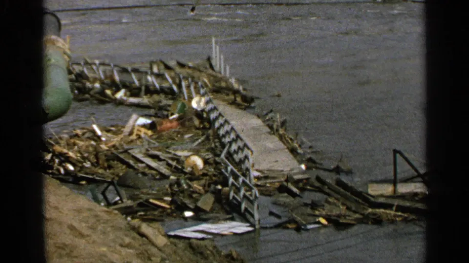 1965 Minnesota waterfront footage showing a damaged dock or walkway, scattered debris, dark water, and a green pipe partly blocking the view.