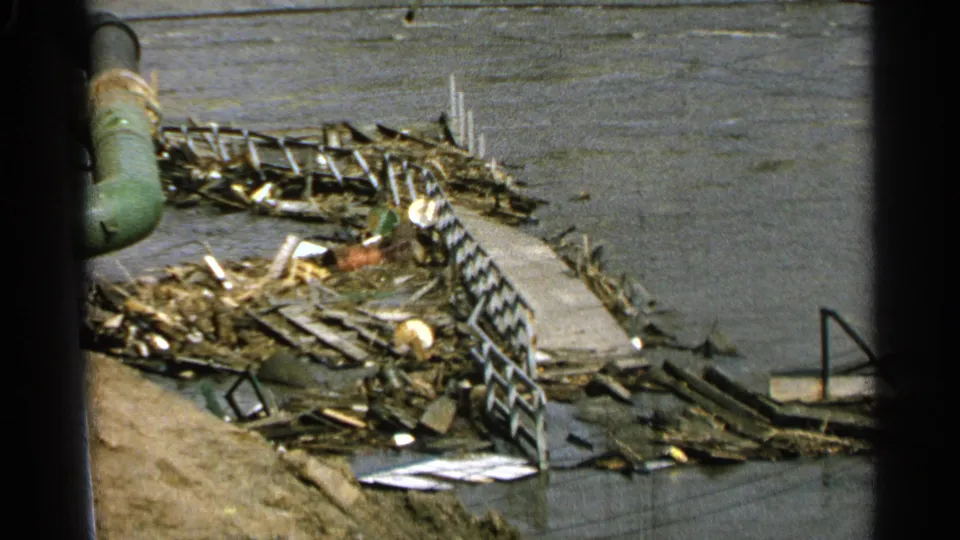 Elevated 8mm view of storm debris around a damaged waterfront dock in Minnesota, with railings, lumber, water, and a green pipe visible.