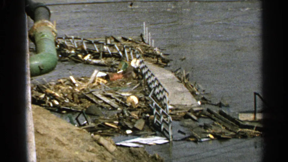 1965 home-movie frame showing an obstructed elevated view of a damaged waterfront walkway with scattered lumber, railings, and water beyond.