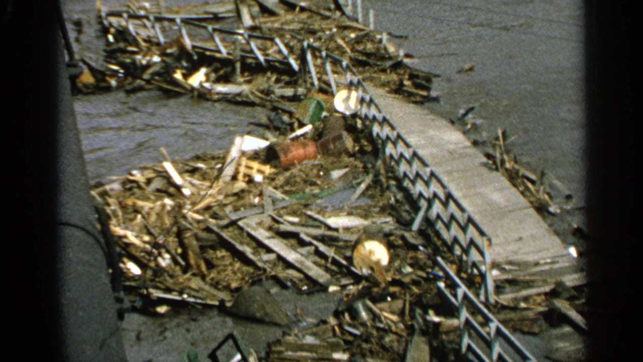 Flood debris and a damaged riverside walkway seen through dark foreground edges.