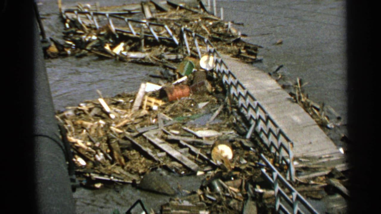 1965 Minnesota home-movie frame showing wreckage, a concrete walkway, and dark river water after flooding.